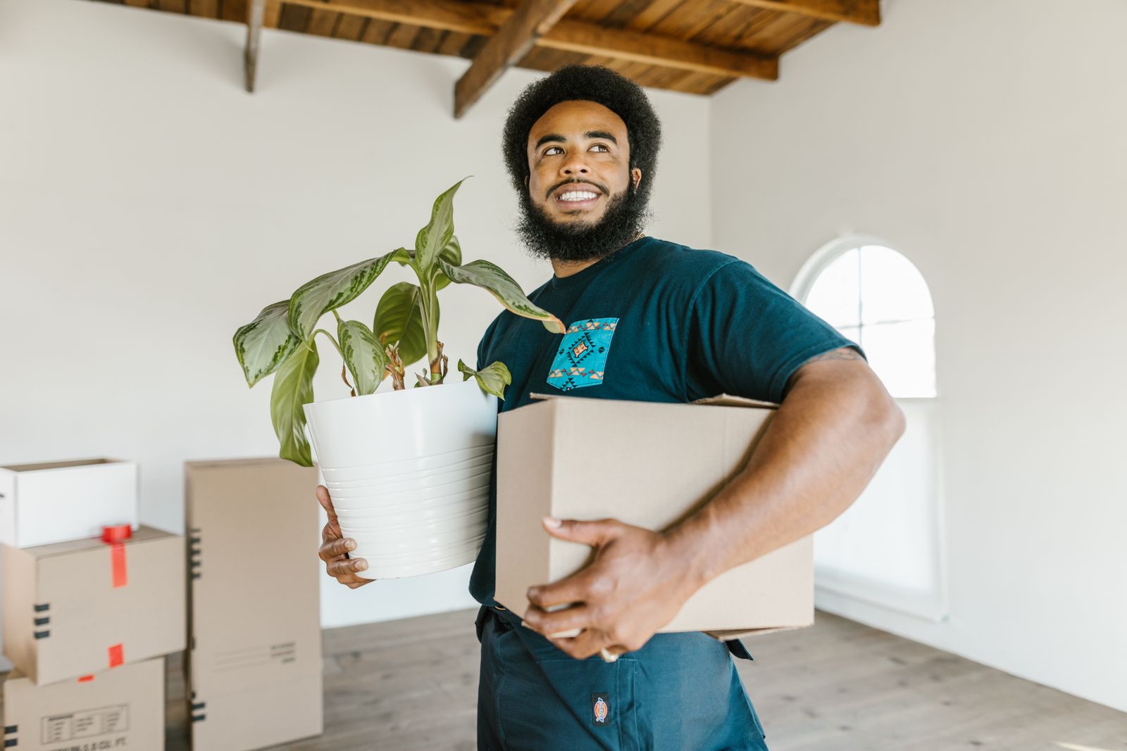 Smiling mover carrying a cardboard box and a potted plant into a new home